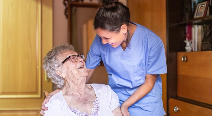 Eine ältere Frau sitzt auf einem Stuhl und schaut lachend zu einer Pflegerin hoch, die hinter dem Stuhl steht und die Seniorin sanft an den Schultern fasst.