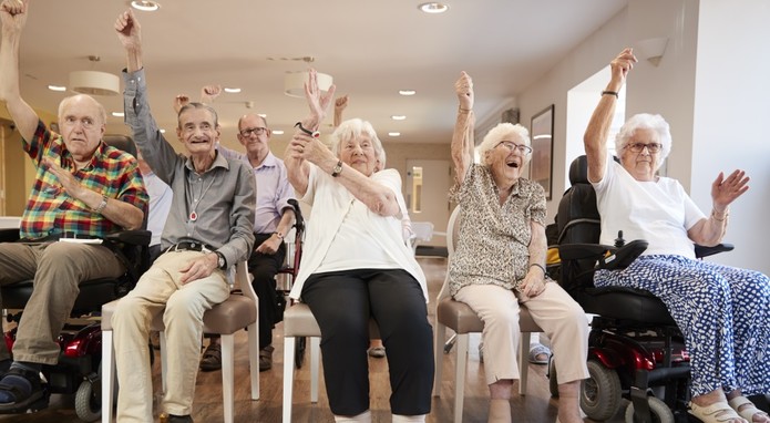 Eine Gruppe Senioren - drei Frau und zwei Männer - beteiligen sich freudig an der Seniorengymnastik. Sie sitzen auf Stühlen und teils im Rollstuhl und recken einen Arm in die Höhe.