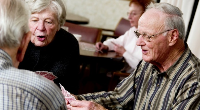 Drei Senioren spielen Karten miteinander. Man sieht eine Frau und einen Mann, die dritte Person sitzt mit dem Rücken zur Kamera.