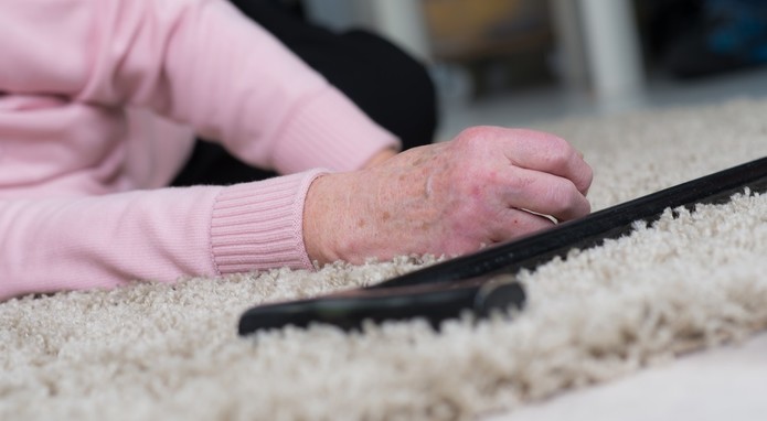 Das Foto zeigt eine ältere Frau, die auf den Teppichboden gestürzt ist. Ein Gehstock liegt neben ihr.