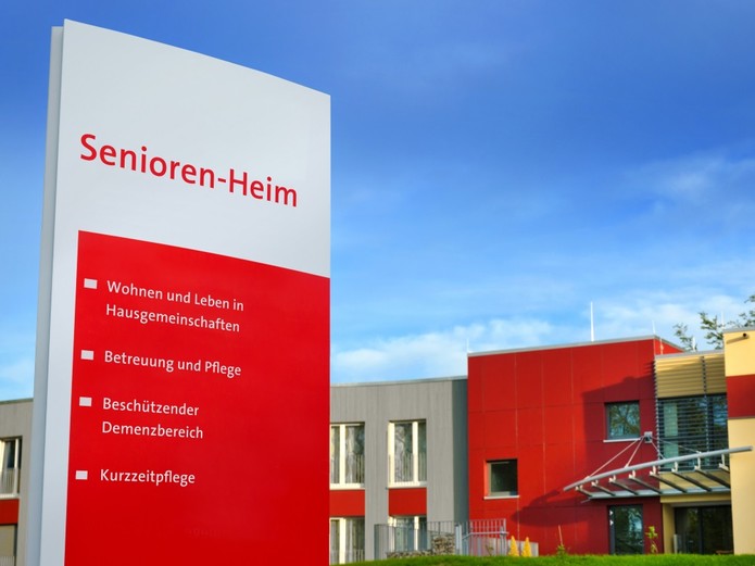 Das Bild zeigt eine Info-Stele, mit der auf die Angebote eines Seniore-Heims hingewiesen wird. Im Hintergrund ist das moderne Pflegeheim mit seiner bunten Fassade zu sehen.